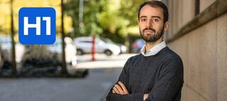 Man in sweater standing with hands crossed next to H1 logo